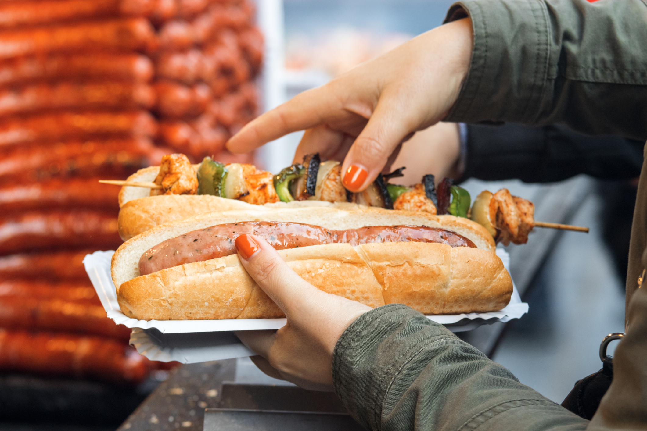 Woman bought her meal at Christmas street market. She holds paper tray in left hand, 2 breads on the tray, one with the sausage and  the other one with kebab skewer. Detail image, focus on 1st bread with sausage.